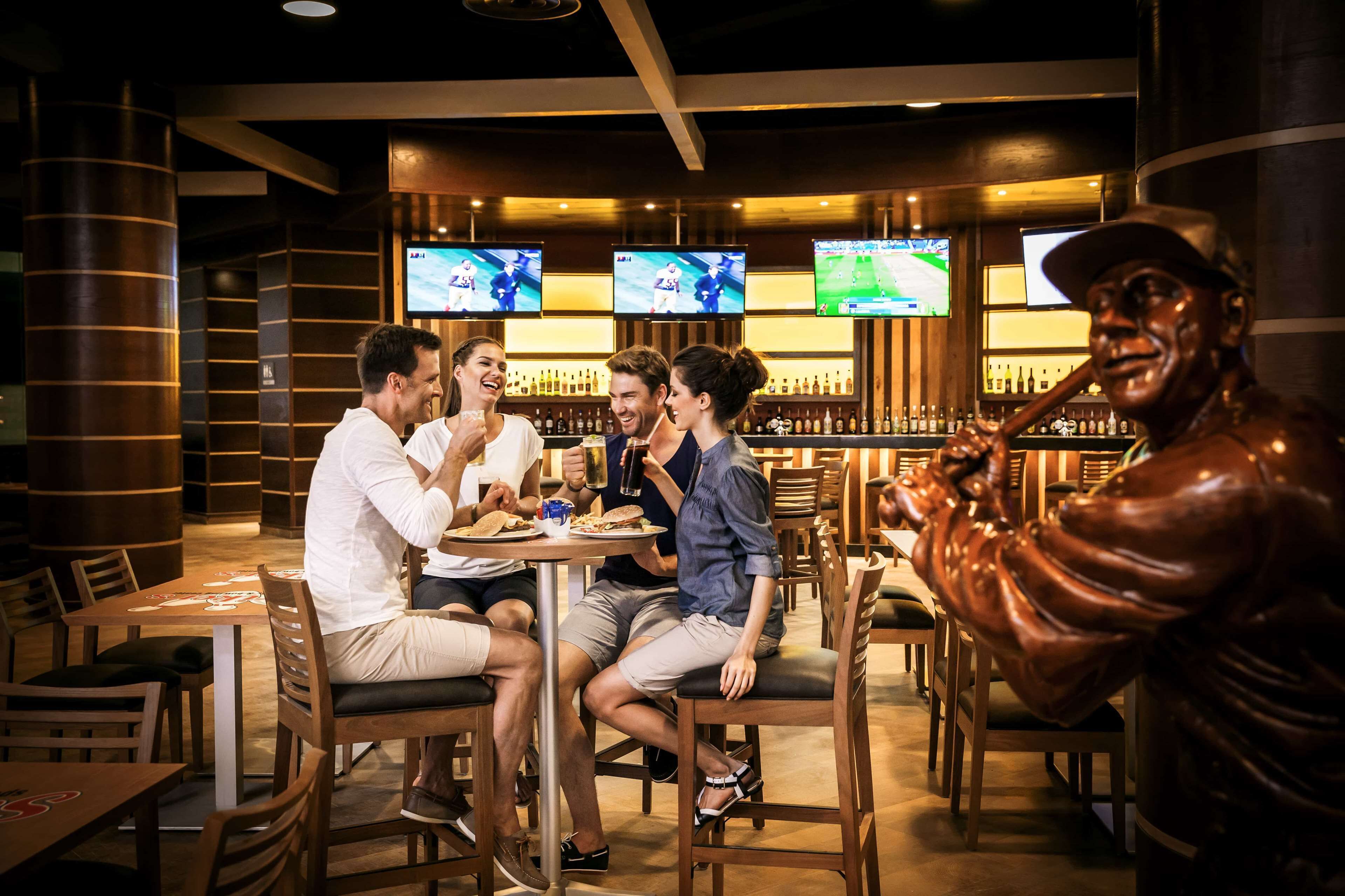 A bar area with multiple screens displaying sports, wooden furniture, and a bronze statue of a man with a hat.