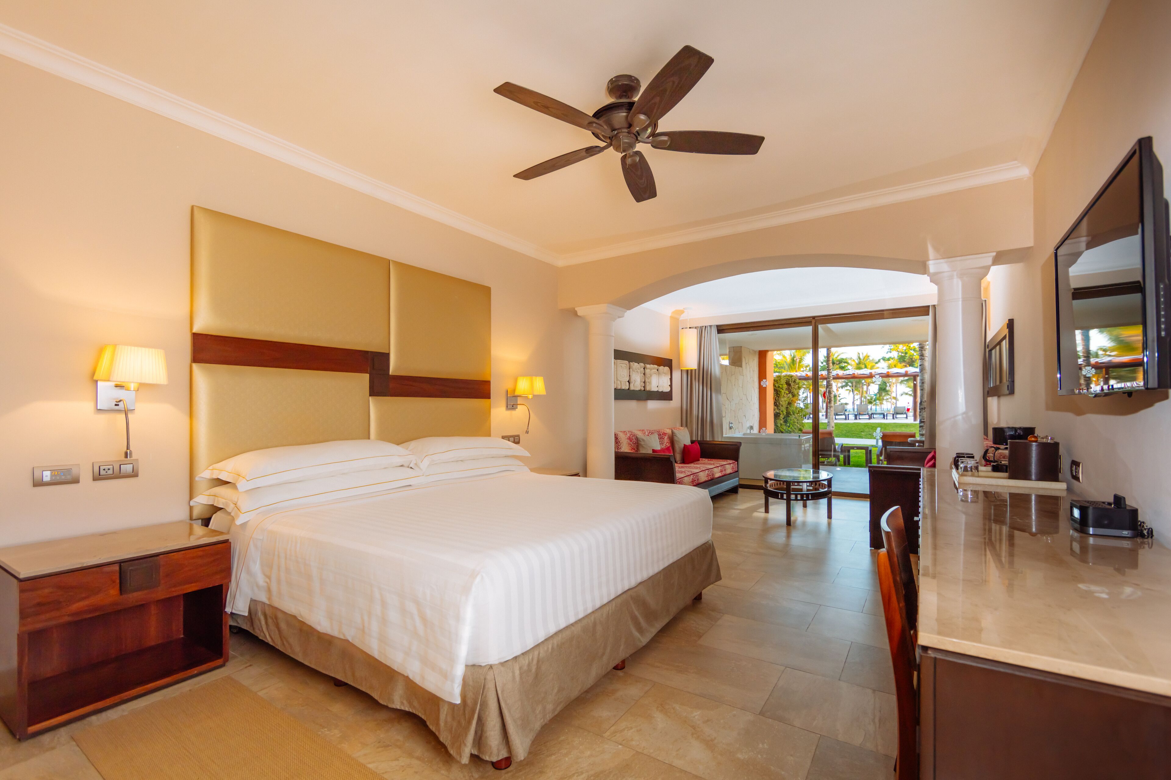 Quarto de hotel com uma cama grande, um ventilador de teto, uma escrivaninha com abajur, uma televisão e vista para a área externa.