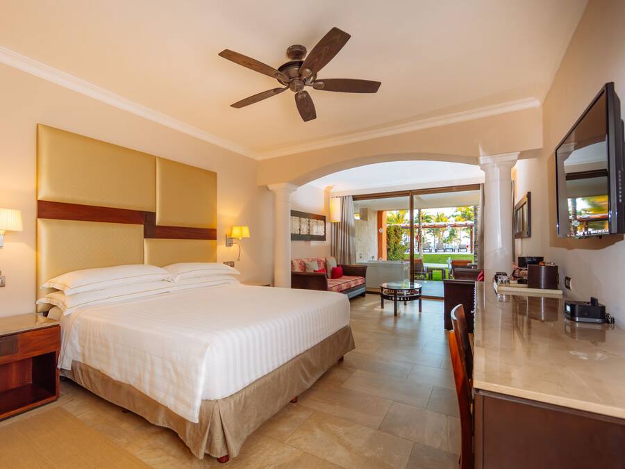 Quarto de hotel com uma cama grande, um ventilador de teto, uma escrivaninha com abajur, uma televisão e vista para a área externa.