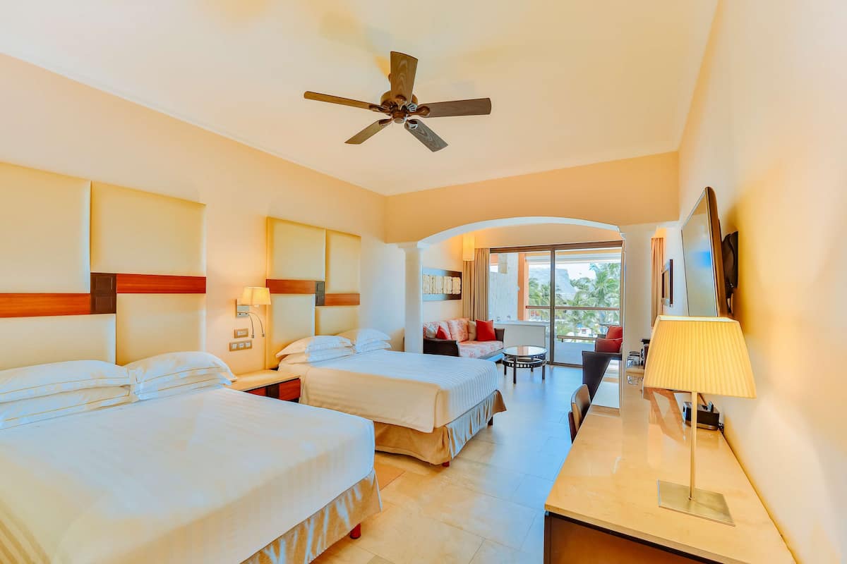 Habitación de hotel con dos camas, un escritorio, una silla, ventilador de techo y vistas al exterior.