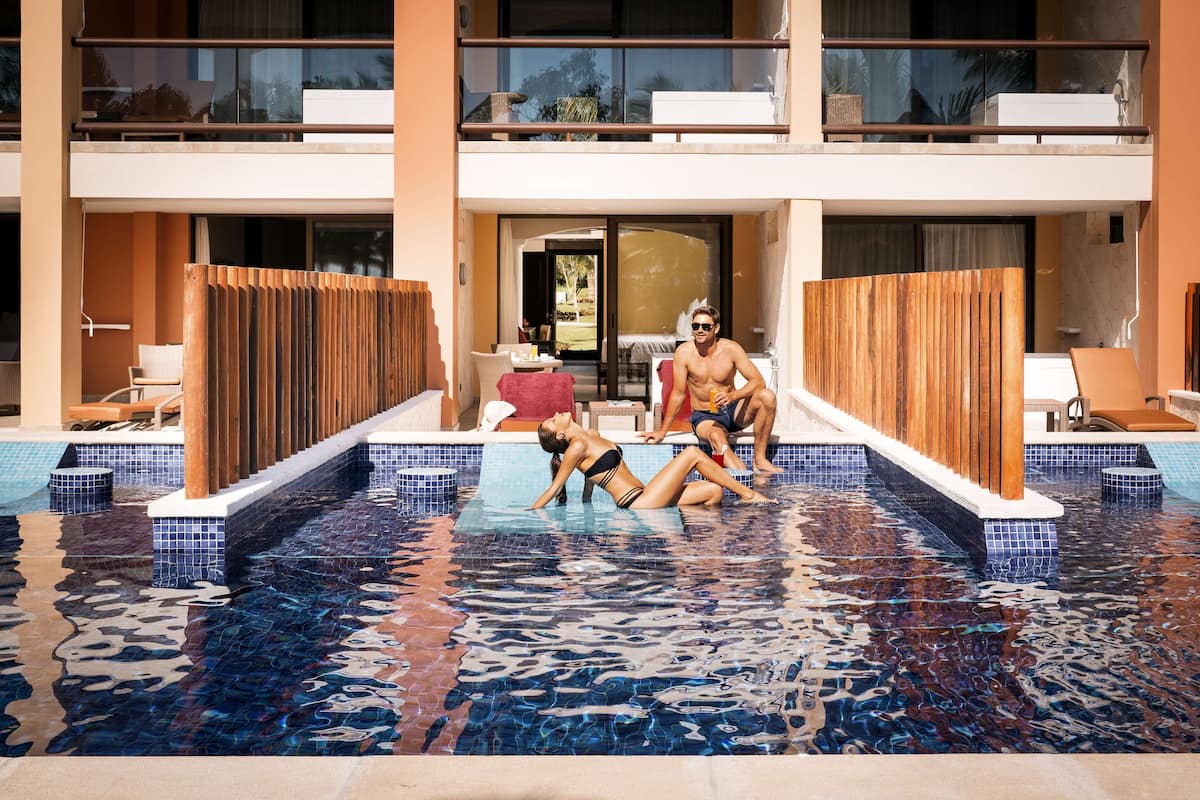 Zona de piscina con terraza de madera, sillas de descanso y vista al interior del edificio.