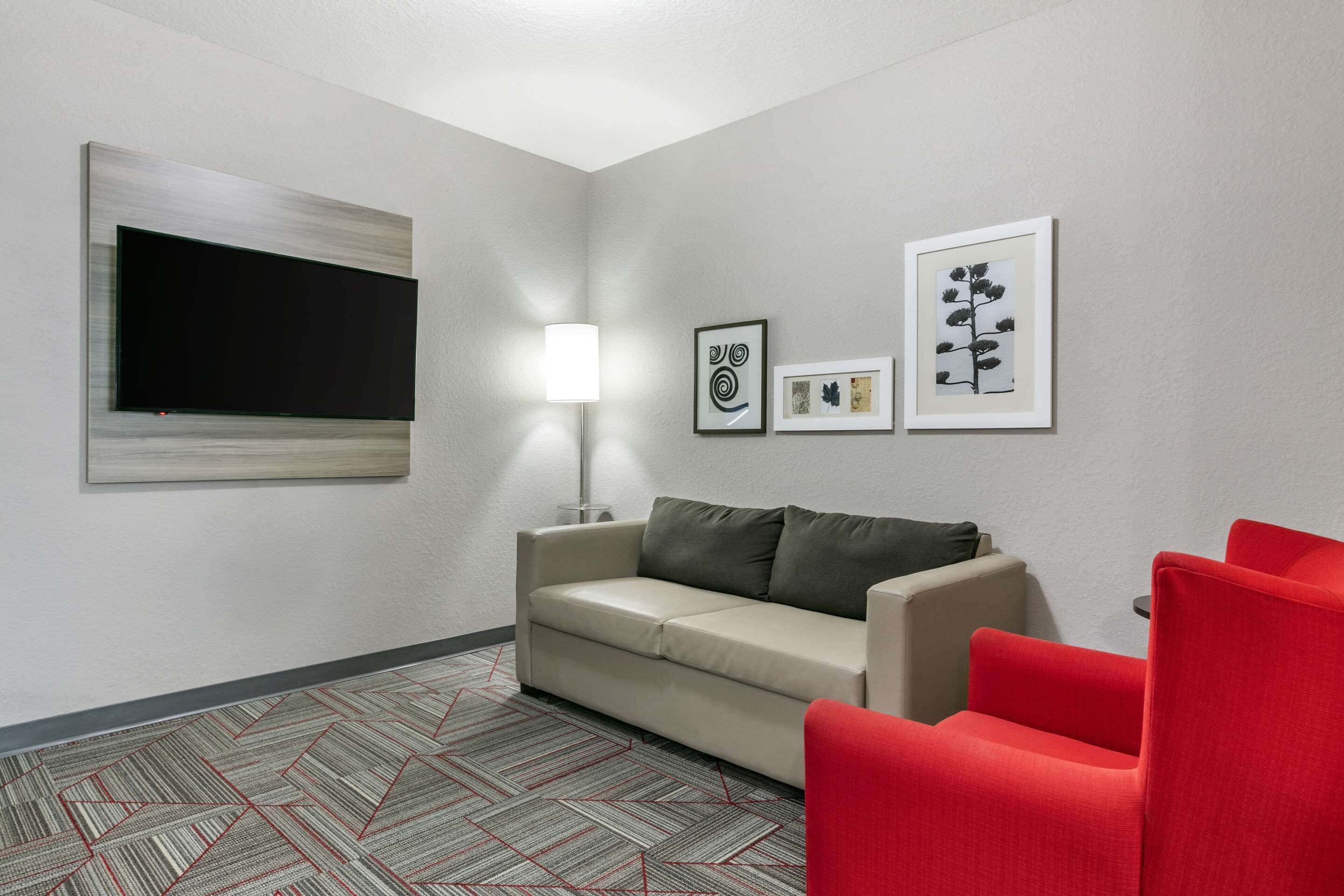 A modern hotel room with a large flat-screen TV mounted on the wall, a sofa, a red chair, and a patterned carpet.