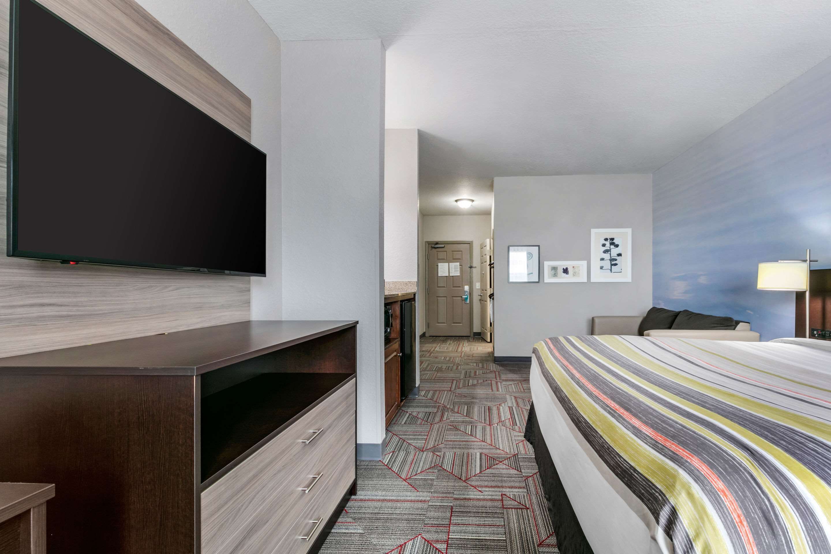 Une chambre d’hôtel dotée d’un grand téléviseur à écran plat fixé au mur, d’un lit avec une couvre-lit à rayures, d’un bureau avec un ordinateur et d’une porte donnant sur une autre pièce.