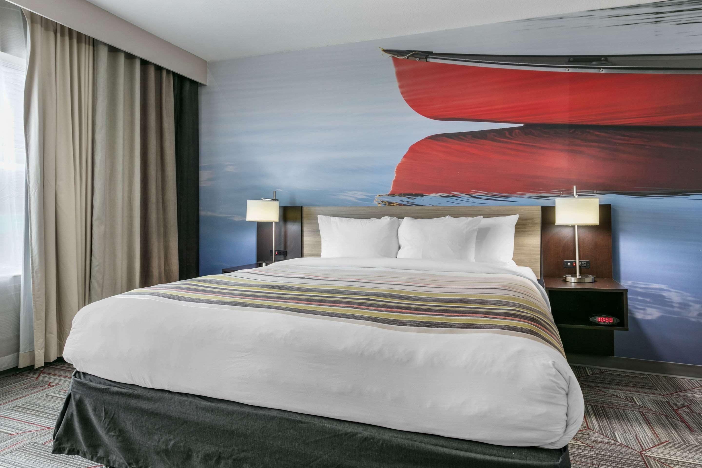 Une chambre d’hôtel avec un grand lit, deux tables de chevet avec des lampes, une horloge numérique et un grand tableau abstrait accroché au mur.