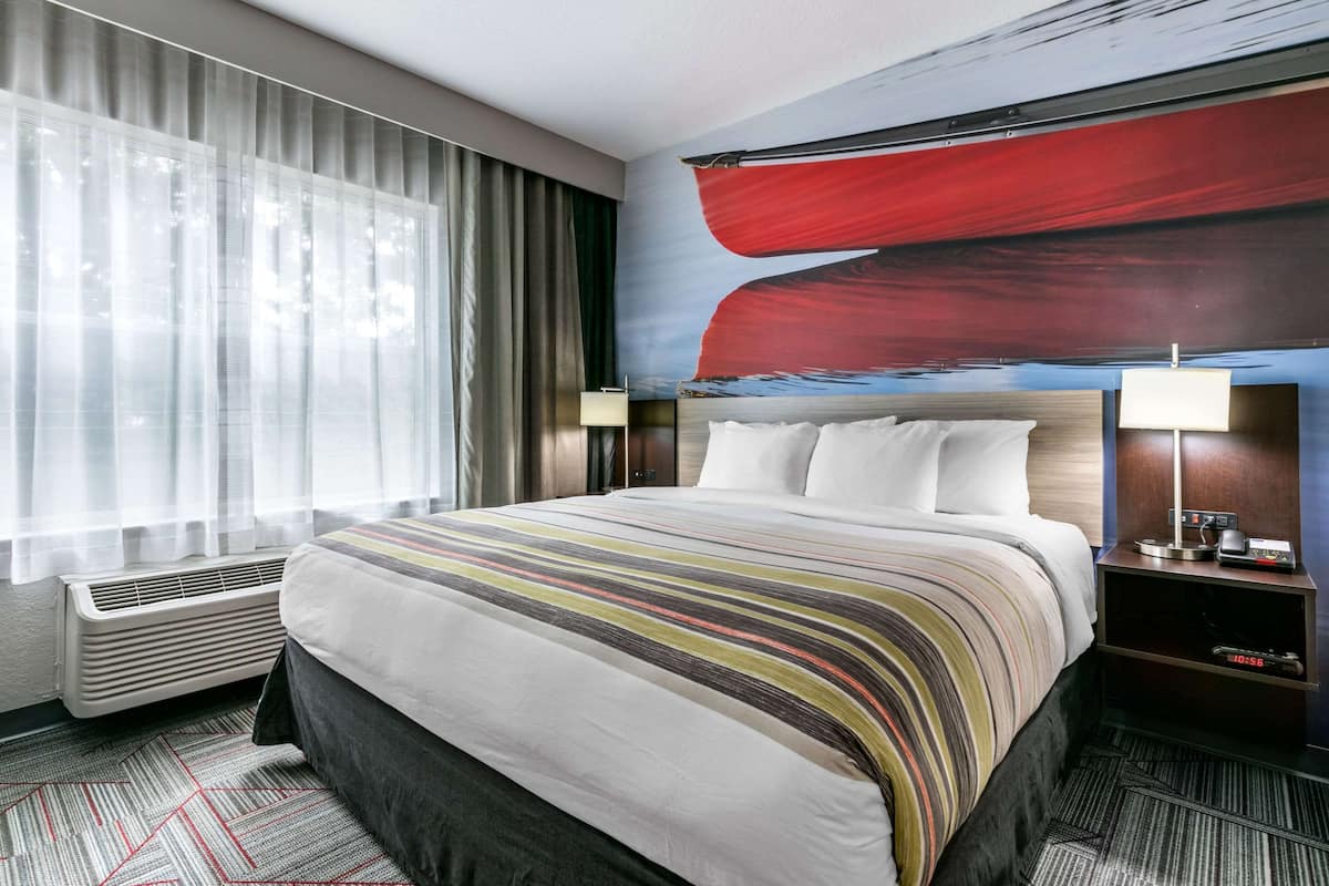 A hotel room with a large bed, a nightstand with a lamp, a window with sheer curtains, and a wall with a red abstract painting.