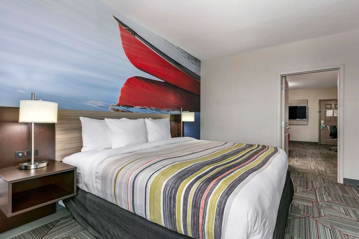 A hotel room with a large bed, two bedside lamps, a nightstand, a painting on the wall, and a view into the hallway.