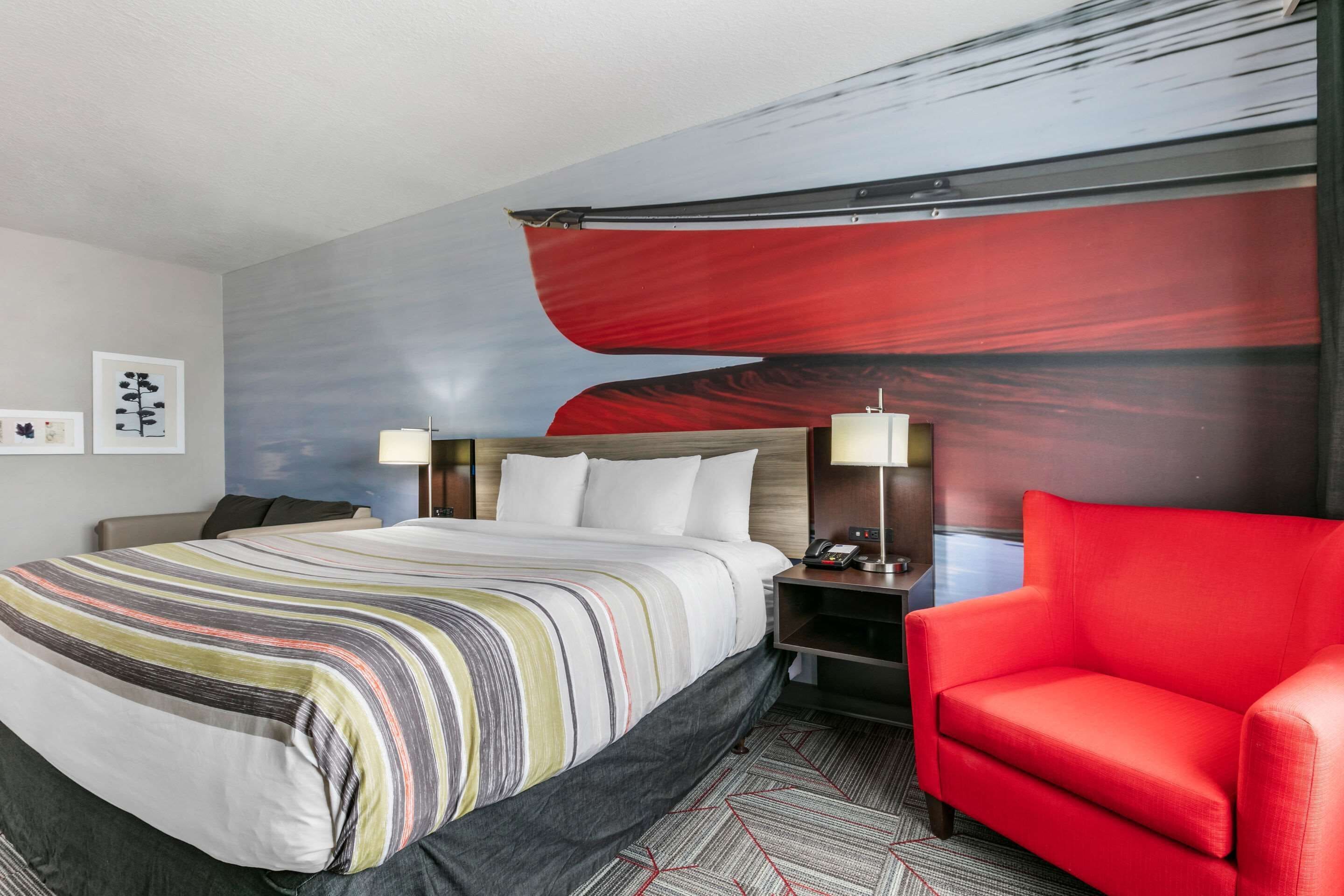 Habitación de hotel con cama, un sillón rojo, una mesita de noche con lámpara y una pared con un cuadro abstracto de gran tamaño.