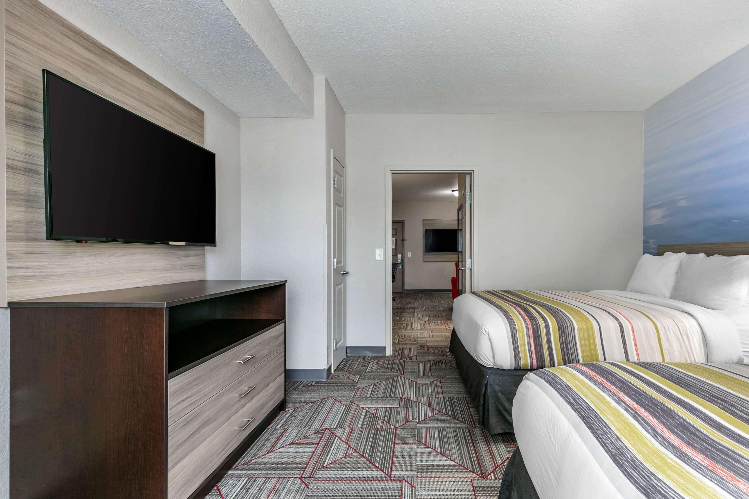 A hotel room with a flat-screen TV mounted on the wall, a bed with striped bedding, a dresser, and a patterned carpet.