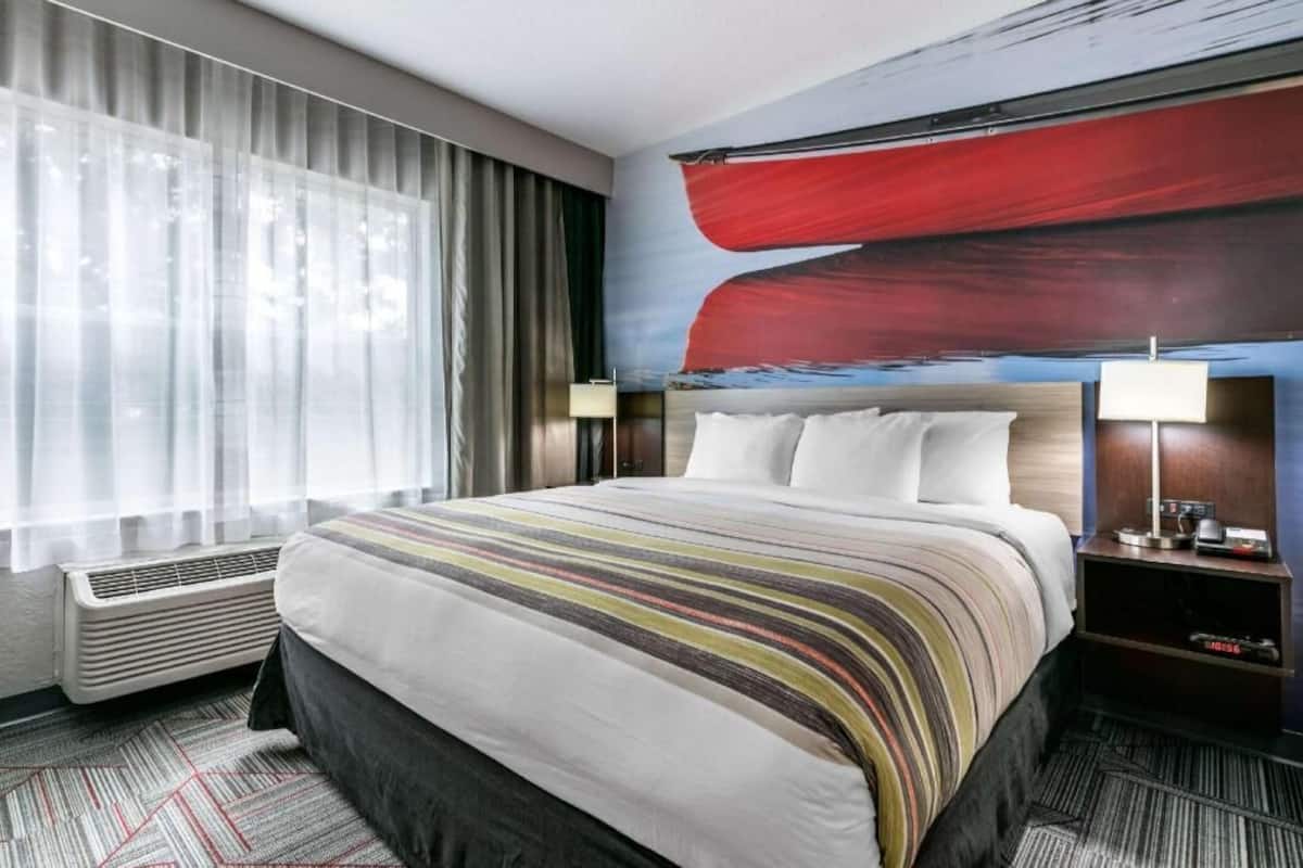 A hotel room with a large bed, a flat-screen TV, a desk with a lamp, and a window with sheer curtains.