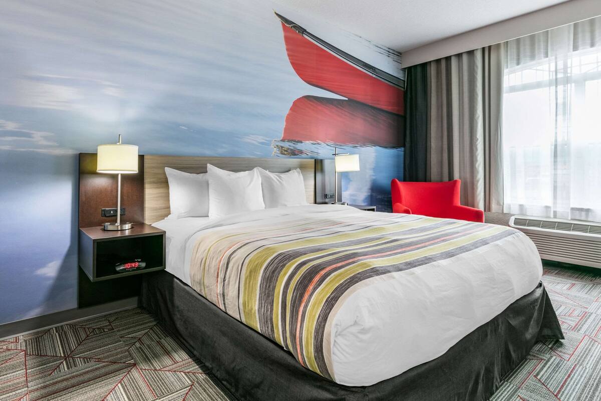 A hotel room with a large bed, a nightstand with a lamp, a red chair, and a large window with curtains.