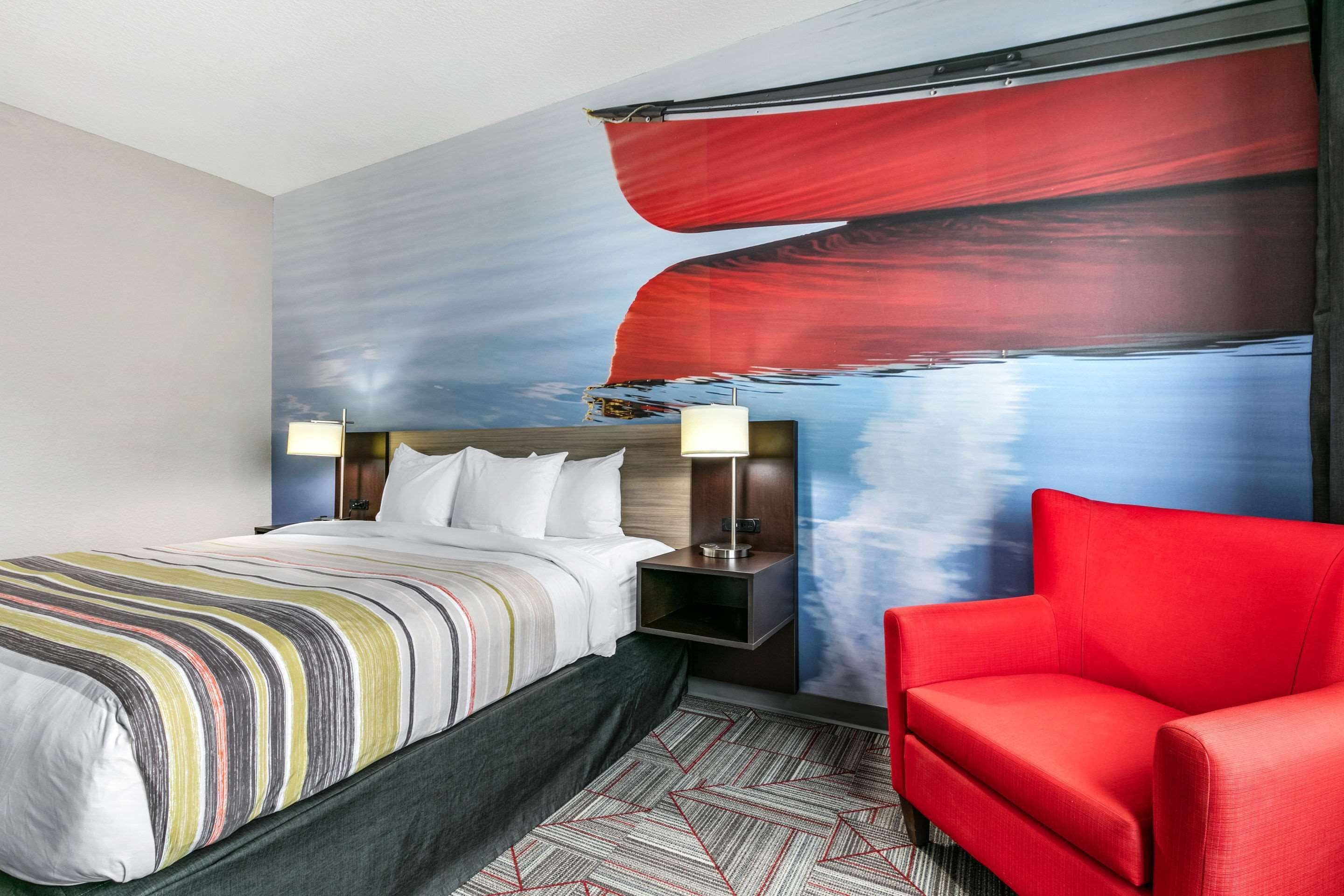 A hotel room with a bed, a red armchair, a nightstand with a lamp, and a wall mural.
