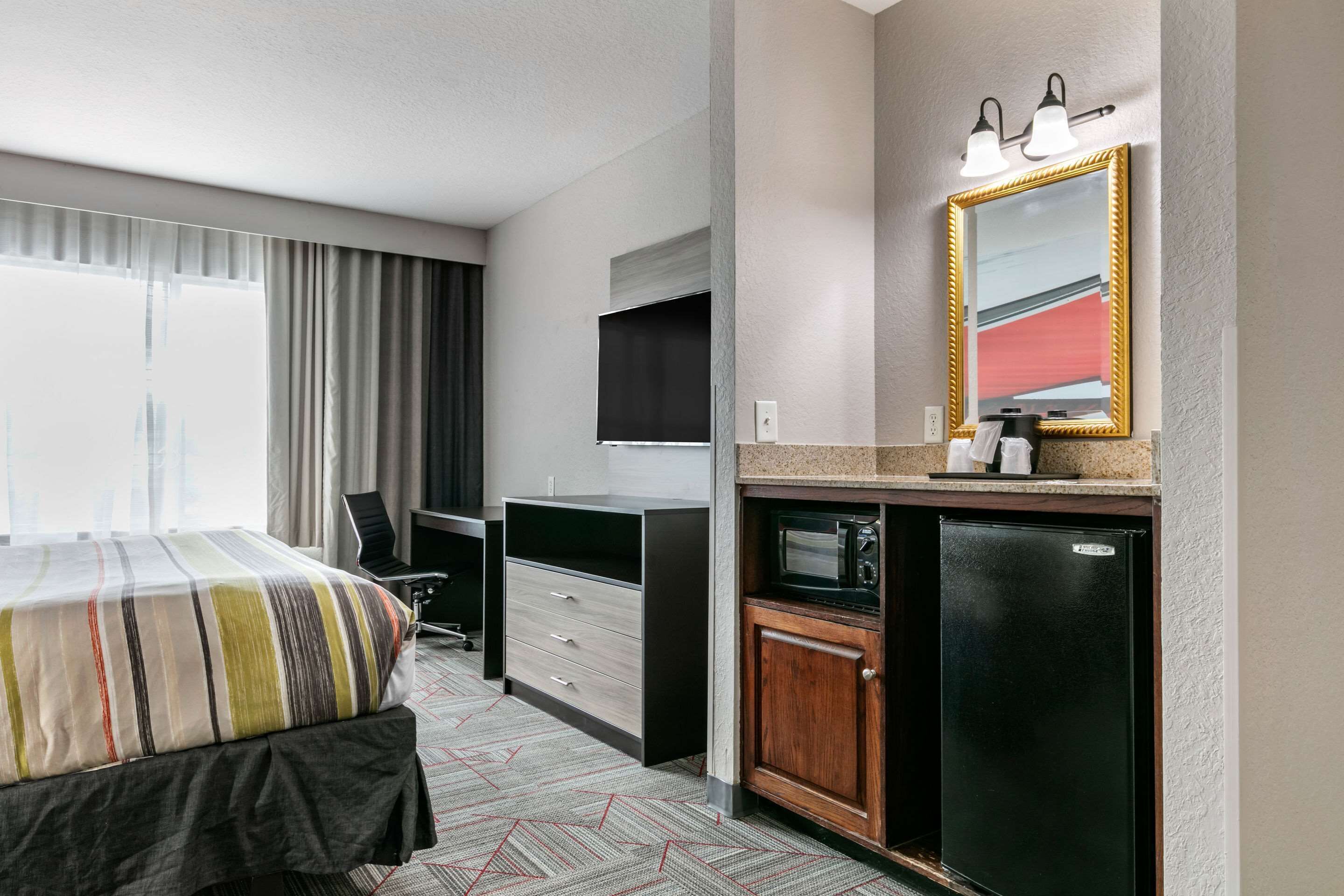 A hotel room with a bed, desk, chair, TV, minibar, and a large mirror.