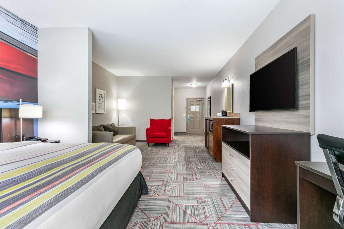 A hotel room with a large bed, a red chair, a desk with a flat-screen TV, and a minibar.