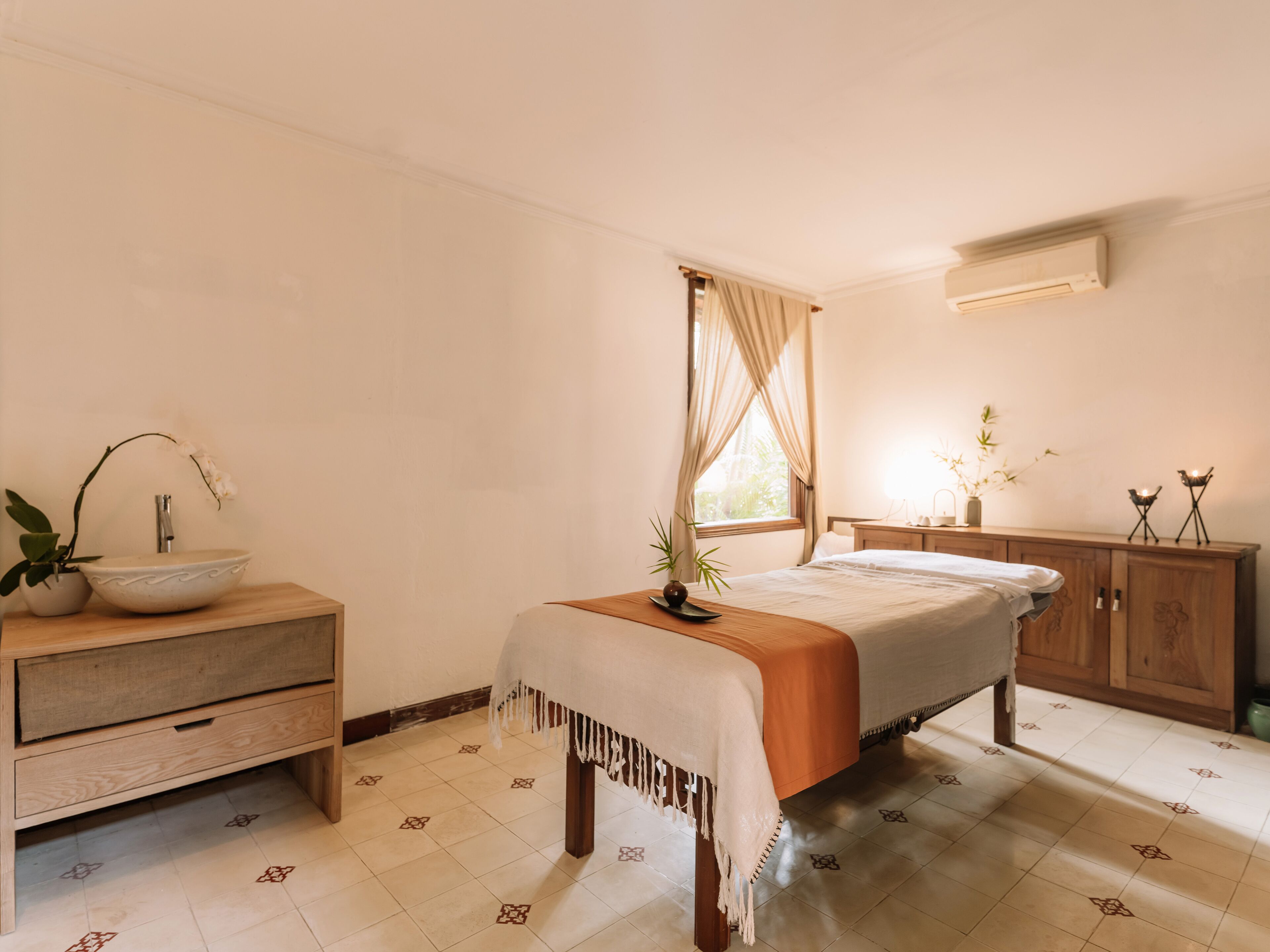 Un dormitorio con cama, mesita de noche de madera, mesita de noche con lámpara, ventana con cortinas y aire acondicionado de pared.