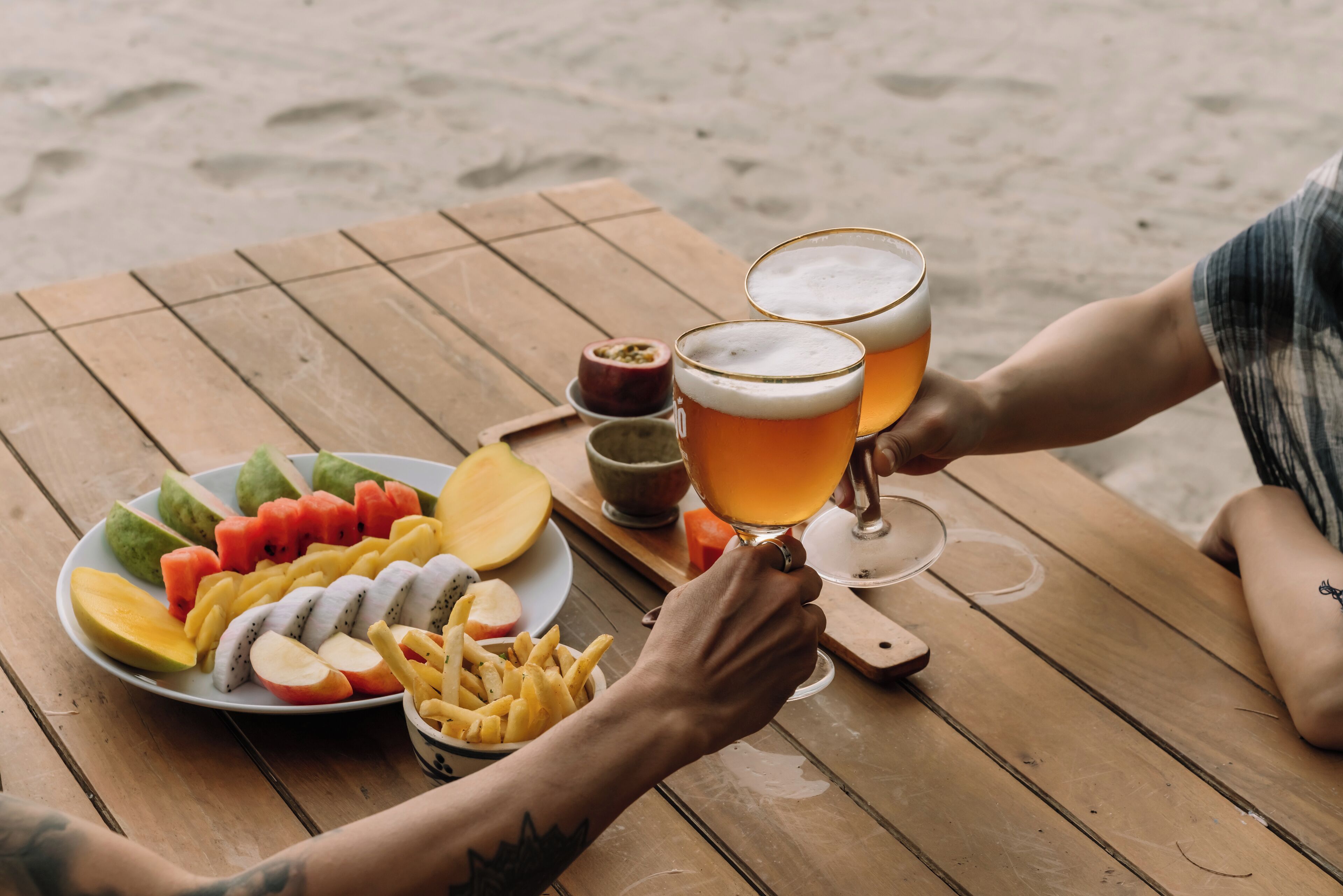 A wooden table with a fruit platter, fries, and two glasses of beer.