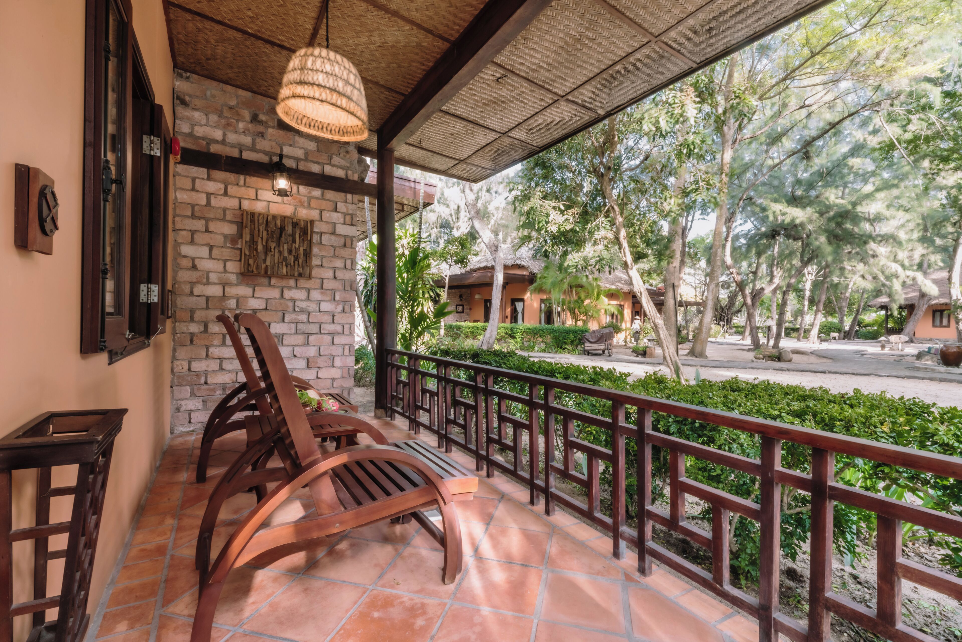 A covered outdoor seating area with wooden chairs and a table, a brick wall, and a view of a landscaped garden and buildings in the background.