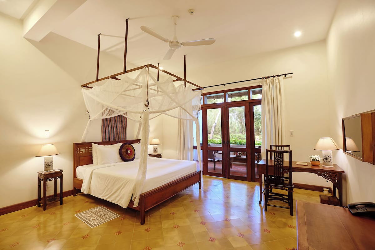 Ein Schlafzimmer mit Himmelbett, Deckenventilator, Schreibtisch mit Stuhl und Blick ins Freie durch eine Glastür.