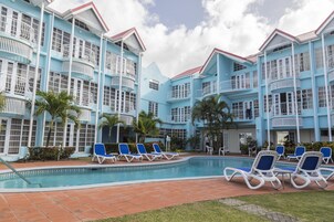 Un hotel con piscina rodeada de sillas reclinables azules, un solárium con baldosas rojas y un edificio de varios pisos con balcones.