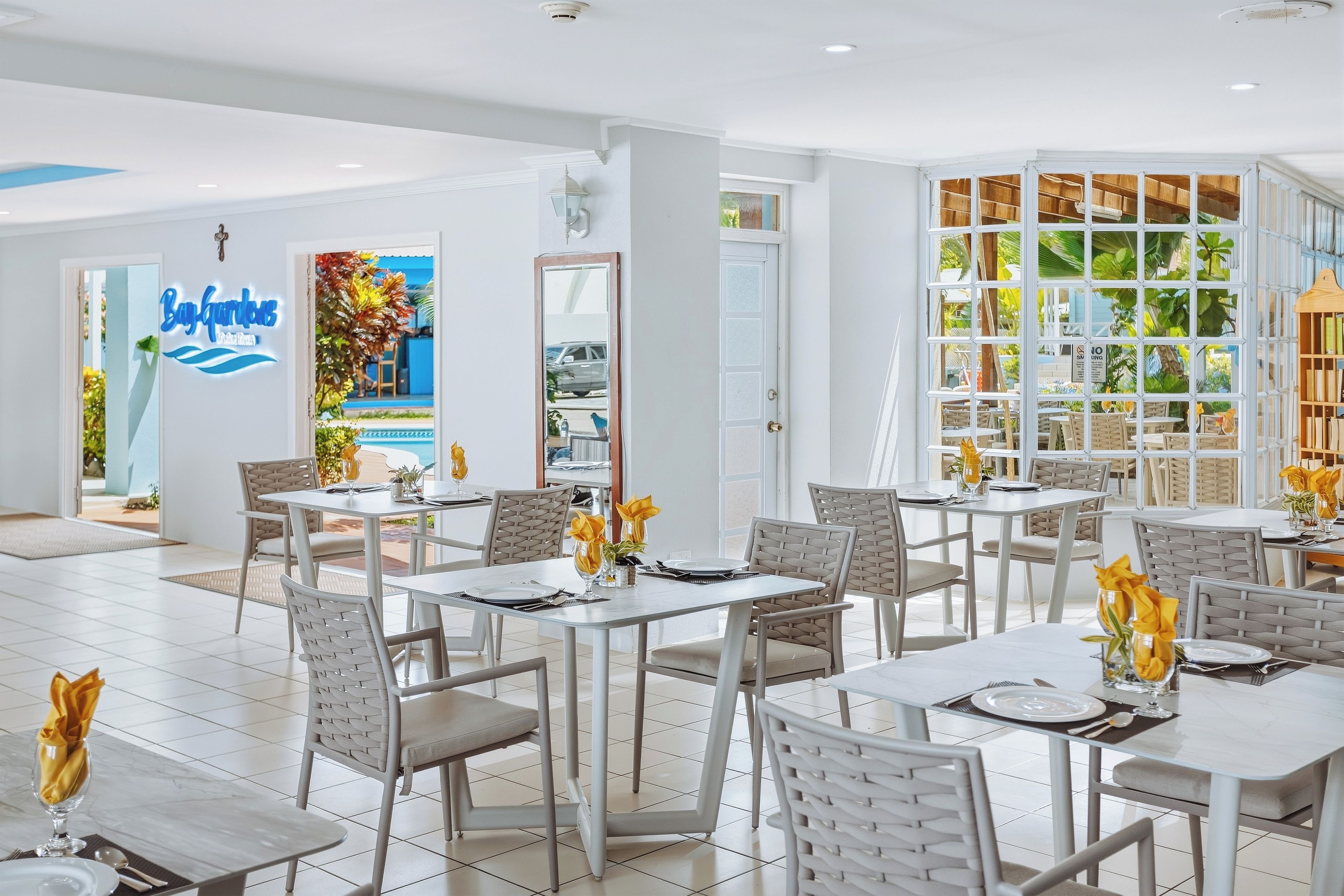 A hotel dining area with tables set for guests, a visible restaurant sign, and a view through a glass partition.