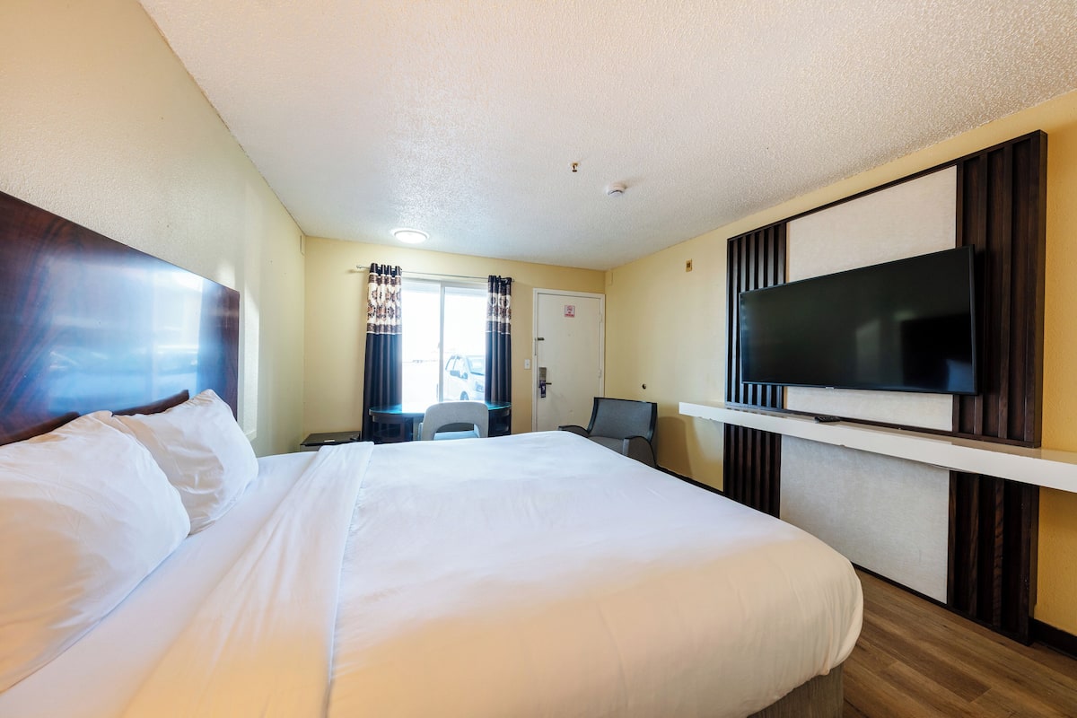 A hotel room with a large bed, a flat-screen TV mounted on the wall, a desk with a chair, and a window with curtains.