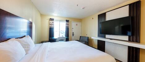 A hotel room with a large bed, a flat-screen TV mounted on the wall, a desk with a chair, and a window with curtains.