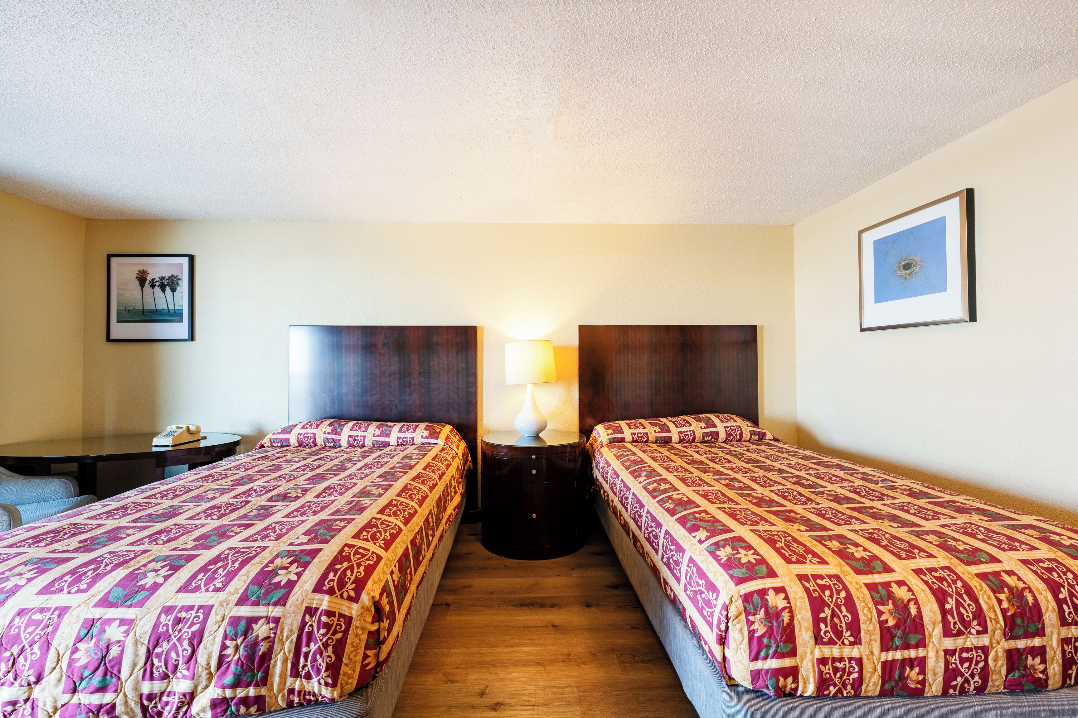 A hotel room with two beds, a small table, a lamp, and framed pictures on the walls.