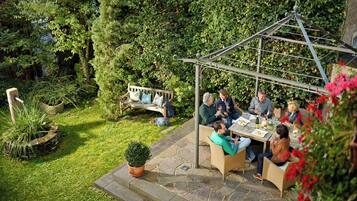 A garden patio with a pergola, stone flooring, and a seating area with wicker furniture.