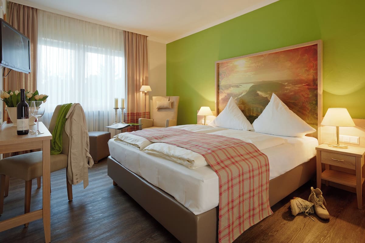 A hotel room with a bed, bedside tables, a desk, a chair, a TV, and a painting on the wall.
