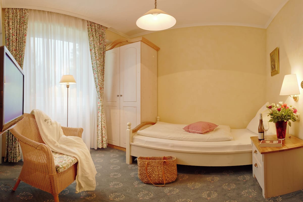 A bedroom with a bed, a chair, a bedside table with a lamp, a television, and a window with curtains.