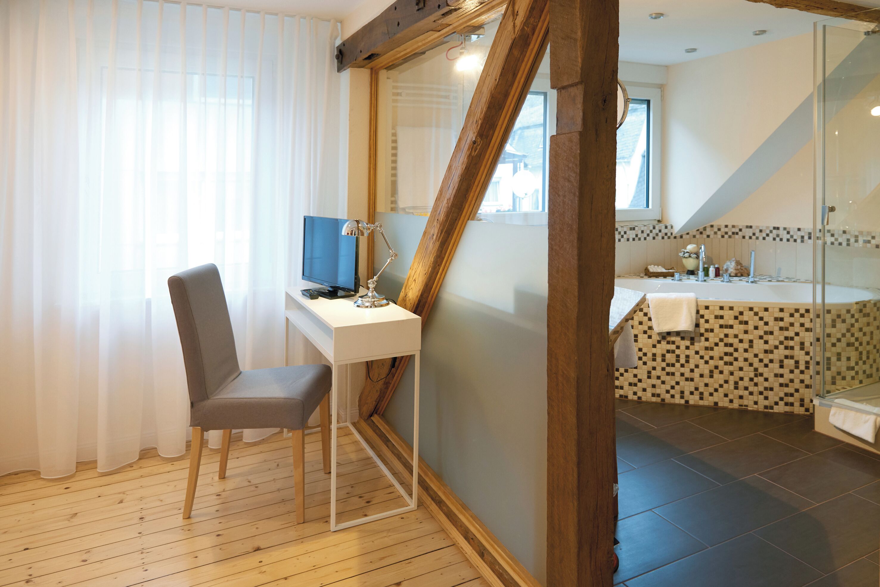 A room with a desk, chair, and a computer. There is a bathroom with a shower and a sink. The room has wooden flooring and a window with curtains.