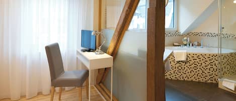Une pièce comprenant un bureau, une chaise et un ordinateur. Elle est également dotée d’une salle de bain avec douche et lavabo. Le sol est en bois et la pièce possède une fenêtre avec des rideaux.