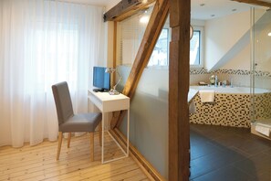 A room with a desk, chair, and a computer. There is a bathroom with a shower and a sink. The room has wooden flooring and a window with curtains.