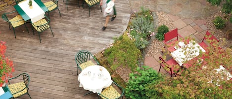Un patio al aire libre con mesas y sillas verdes y rojas, rodeado por un jardín con diversas plantas y un sendero de piedra.