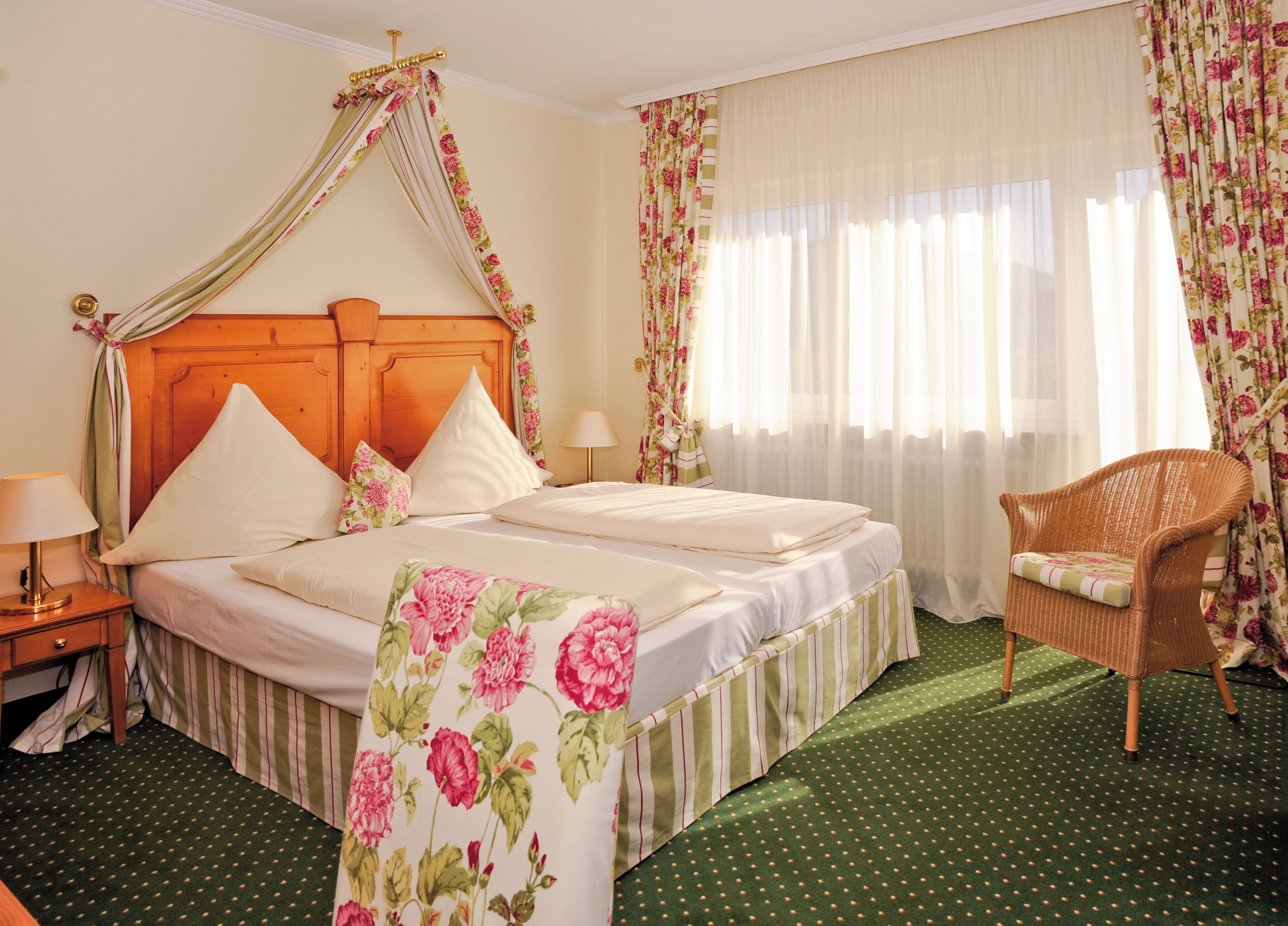 Una camera d'albergo con una testiera in legno, biancheria da letto floreale, un tappeto verde a pois e una sedia in vimini.