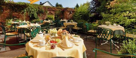 Un espace de restauration en plein air avec des tables rondes recouvertes de nappes beiges, des chaises en métal vert et une variété de plats et de boissons disposés sur les tables.
