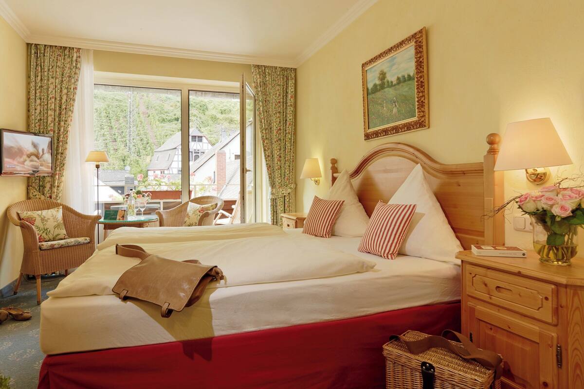 A hotel room with a large bed, a wooden nightstand, a wicker chair, a table with a vase of flowers, and a painting on the wall.