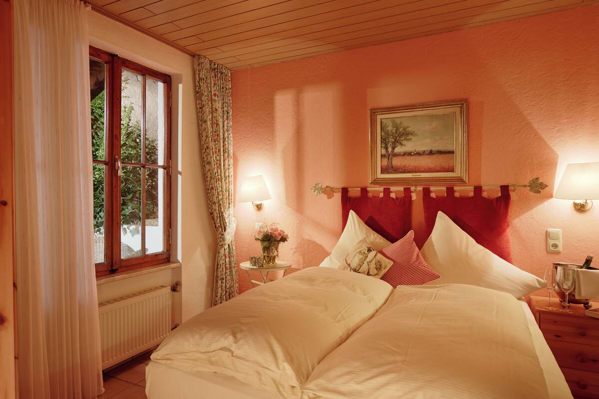 A bedroom with a bed, a window with curtains, a bedside table with a lamp, and a painting on the wall.