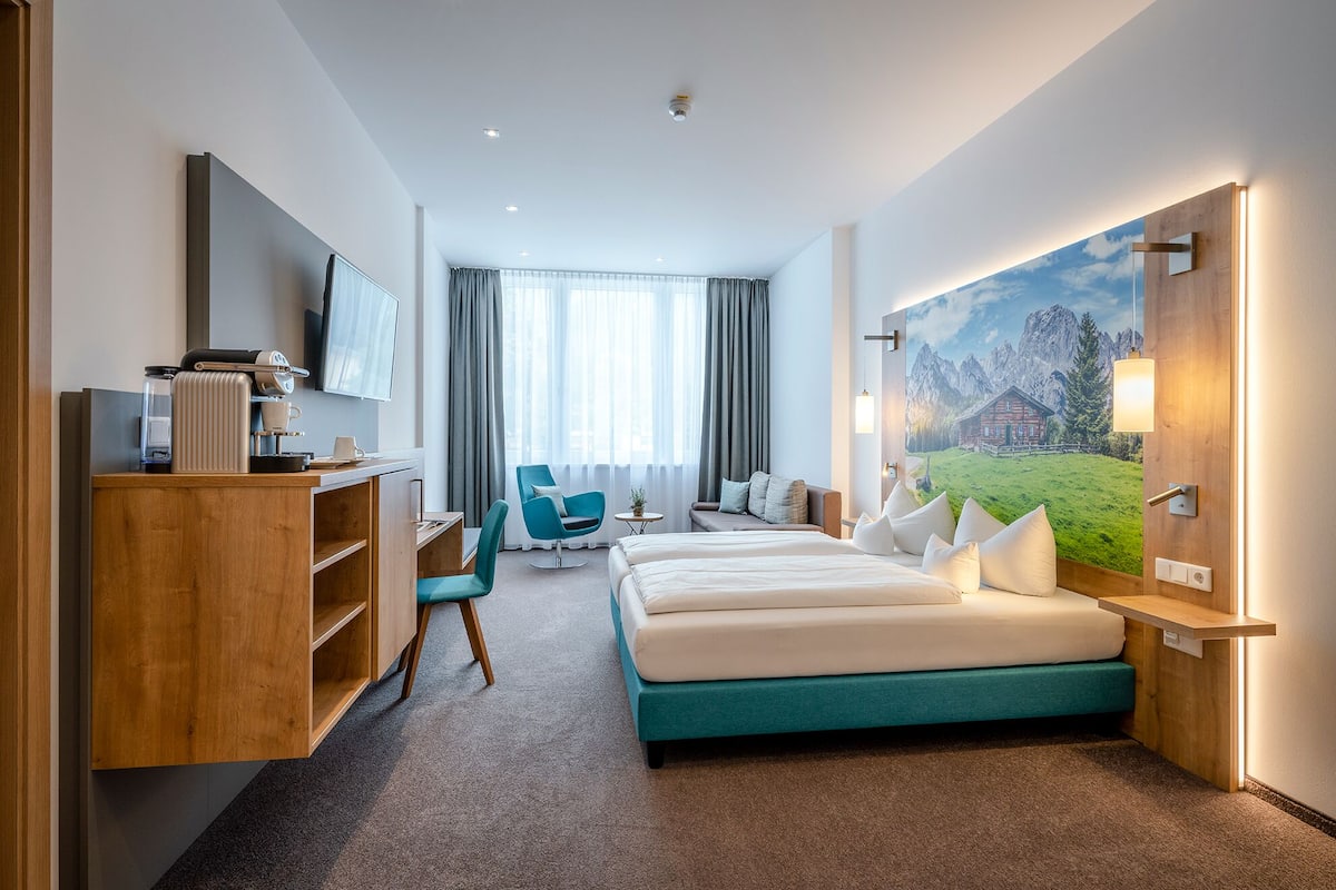 A hotel room with a bed, a desk, a chair, a TV, and a painting of a mountain landscape.