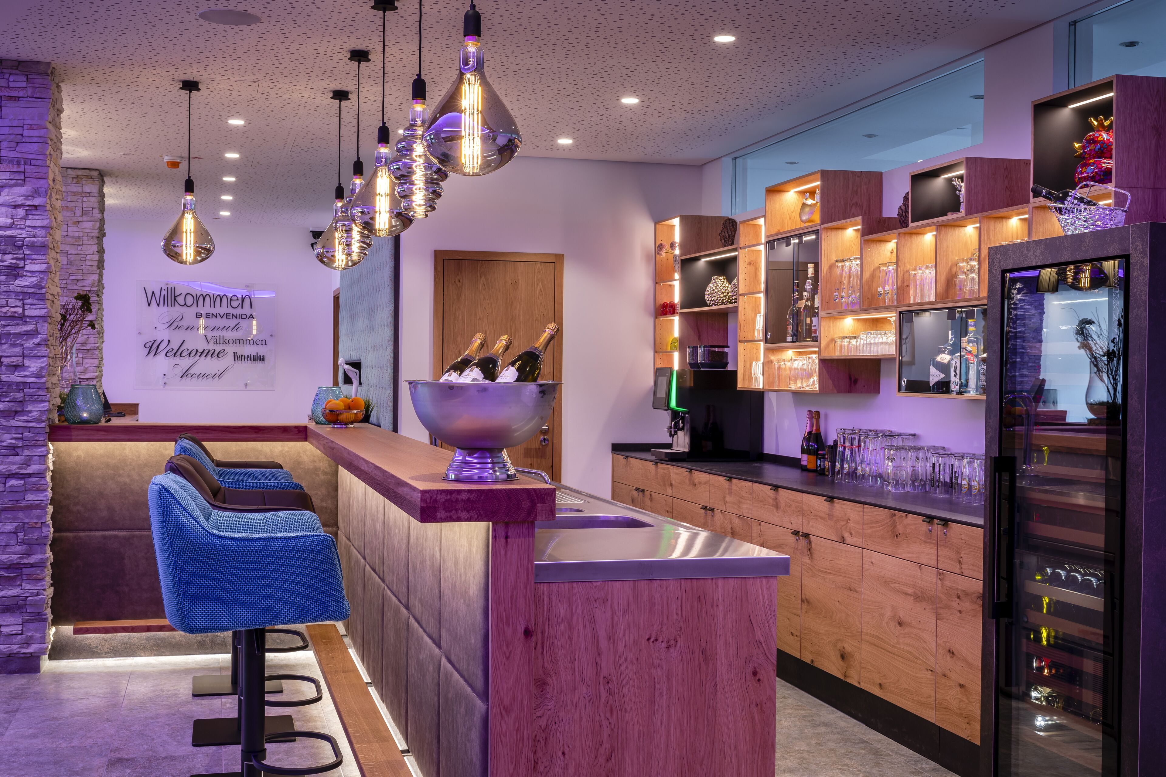 A modern kitchen with wooden cabinets, a bar area with blue bar stools, and a wine fridge.
