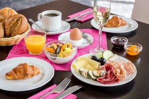 A breakfast setting with croissants, a croissant on a plate, a bowl of fruit, a glass of orange juice, a glass of white wine, and a plate with cheese, olives, and meats.