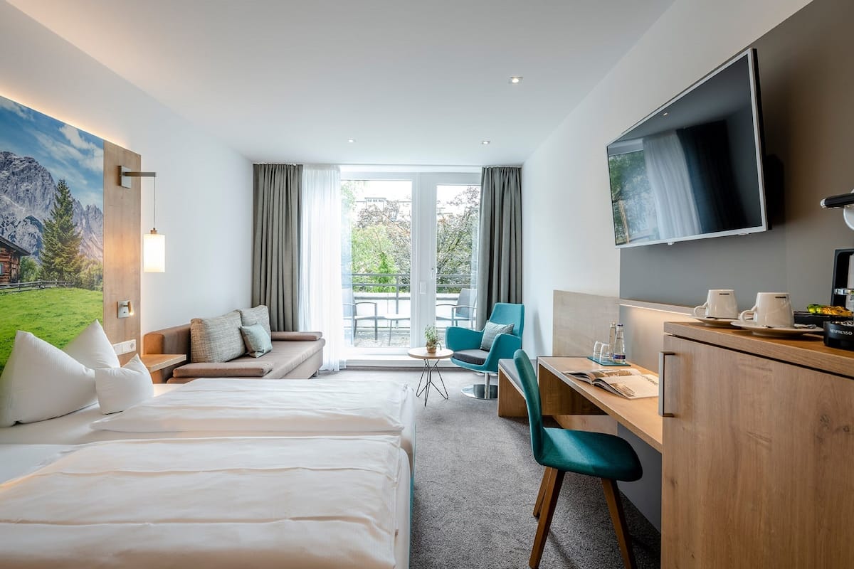 A hotel room with a bed, a desk, a TV, a balcony with a view of the mountains, and a painting on the wall.