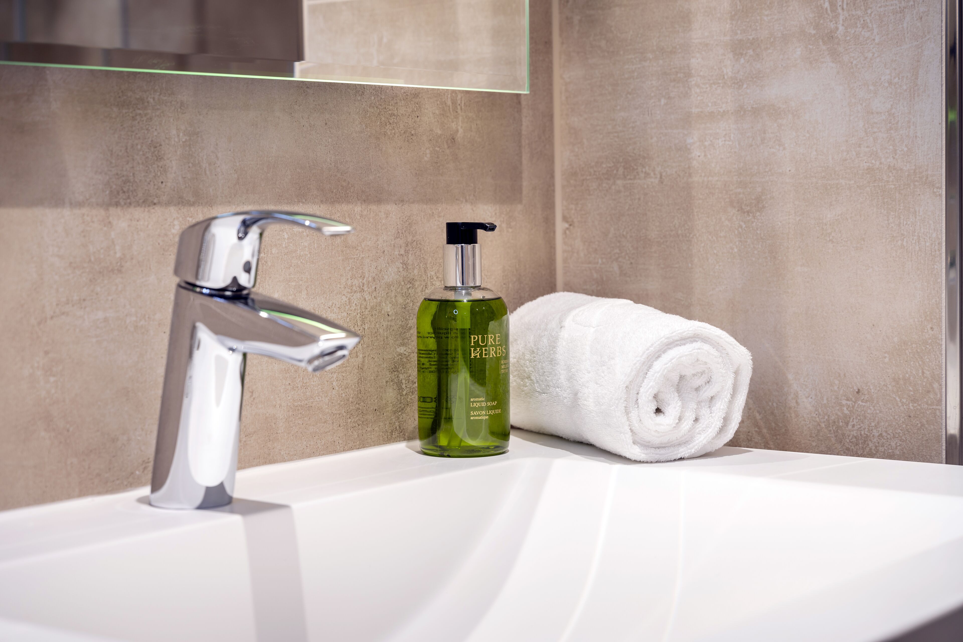 Un baño con un grifo cromado, una botella verde de jabón líquido Pure Herbs y una toalla blanca enrollada sobre un lavamanos blanco.