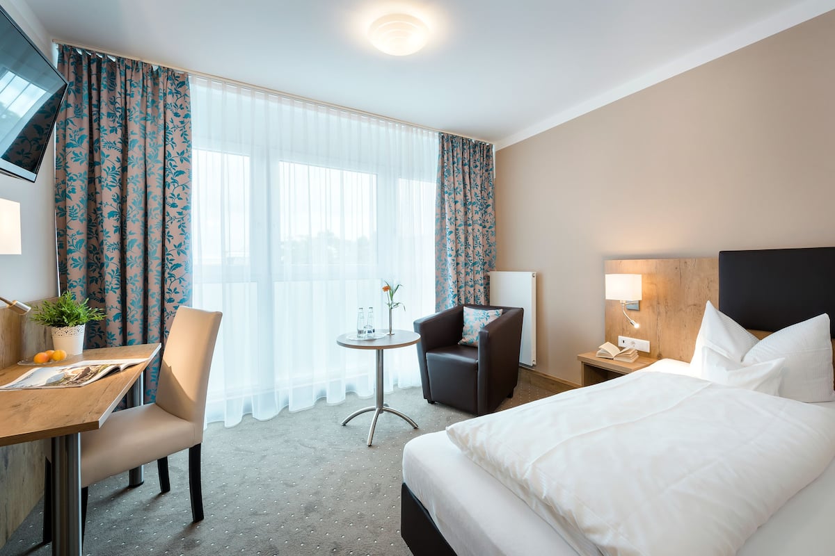 A hotel room with a bed, a desk with a chair, a small table with a plant, and a nightstand with a lamp.