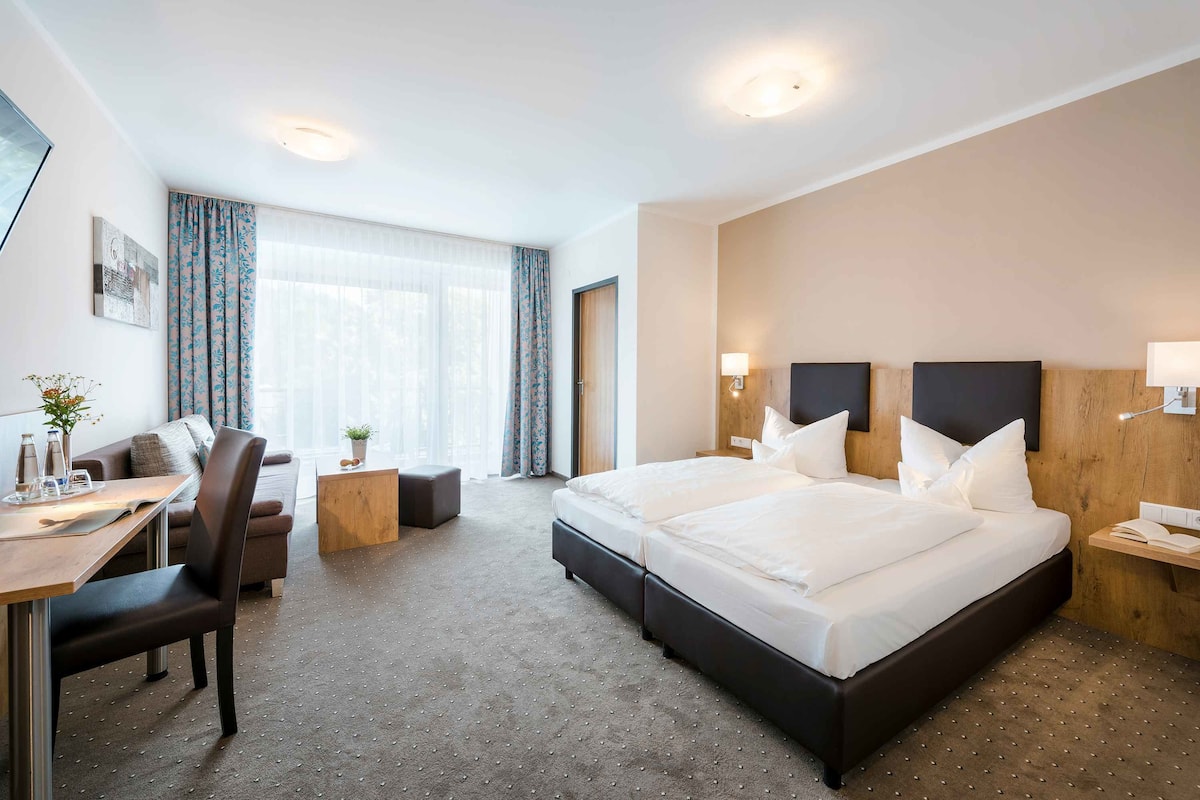 A hotel room with a large bed, a desk with a chair, a small table, and a window with curtains.