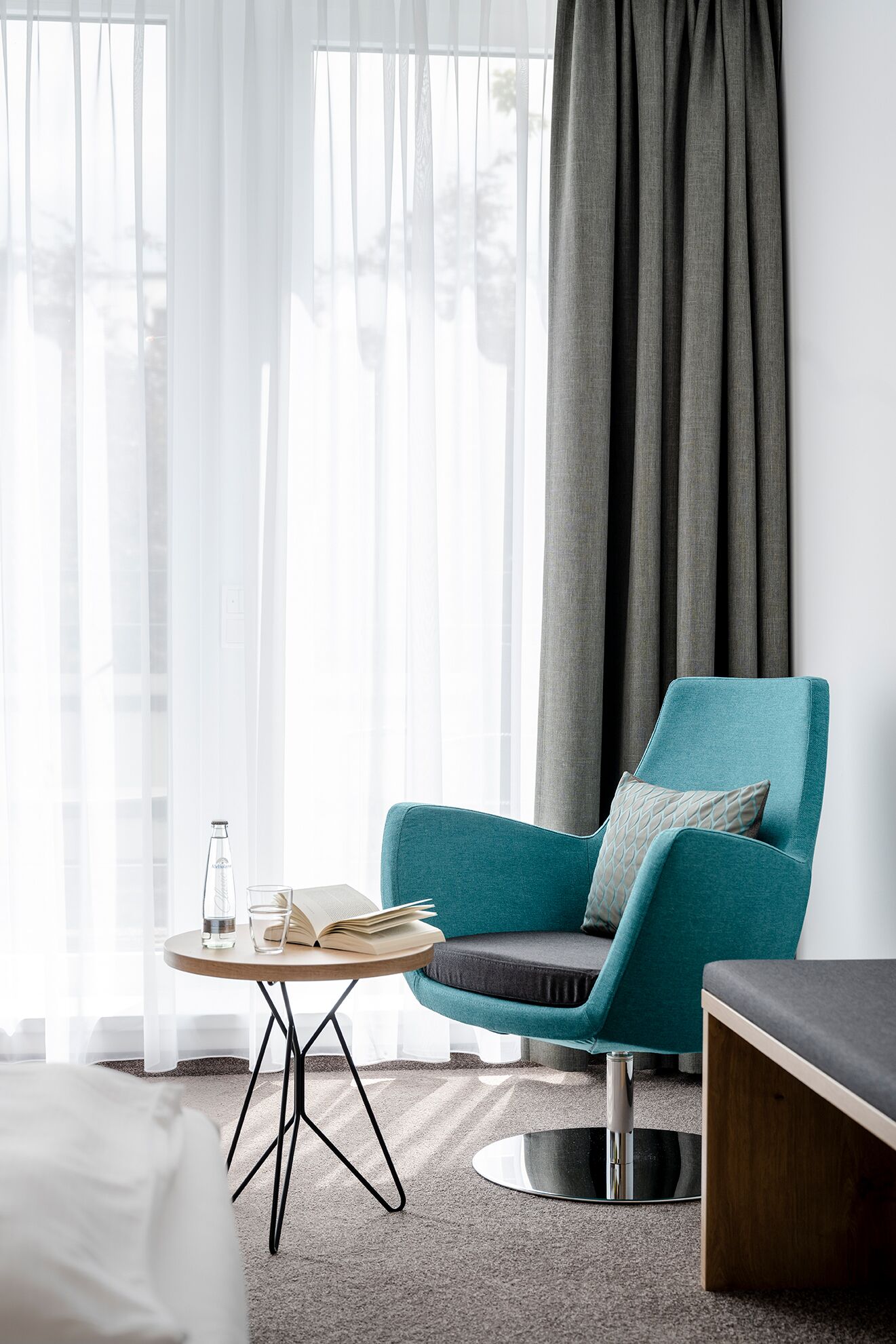 Ein modernes Hotelzimmer mit einem petrolfarbenen Sessel, einem runden Couchtisch und einem Bett.