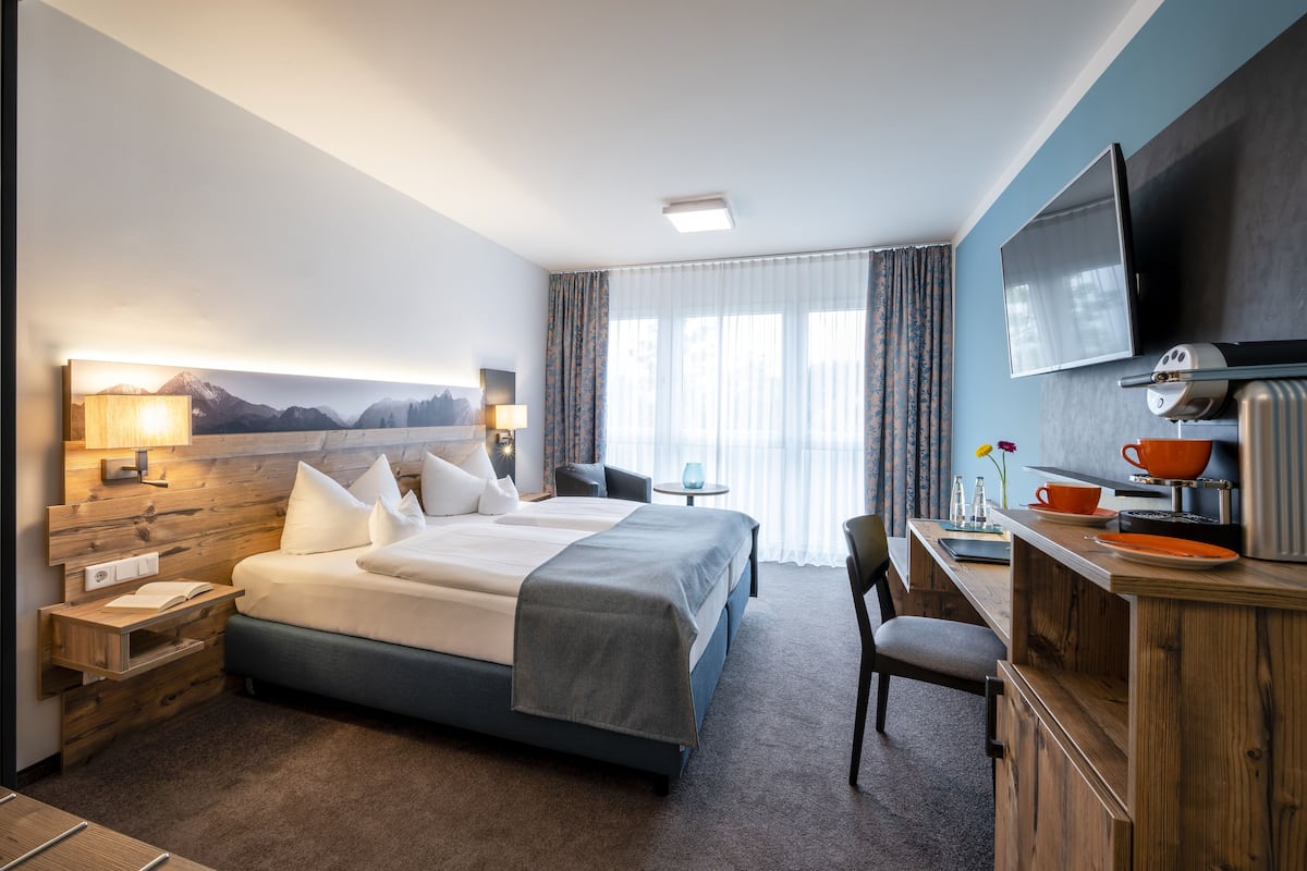 A modern hotel room with a large bed, a desk with a flat-screen TV, and a view of mountains through the window.