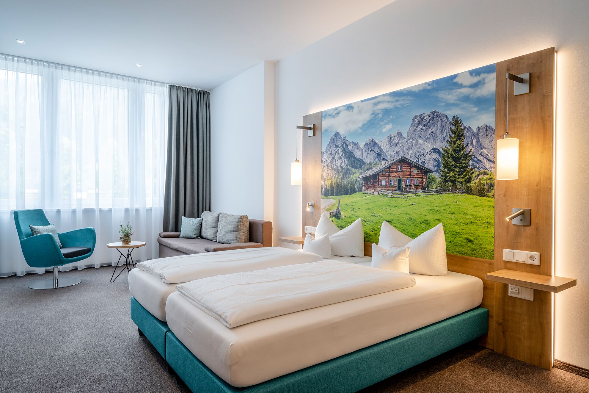 A hotel room with a bed, two chairs, a small table, and a large painting of a mountain landscape.