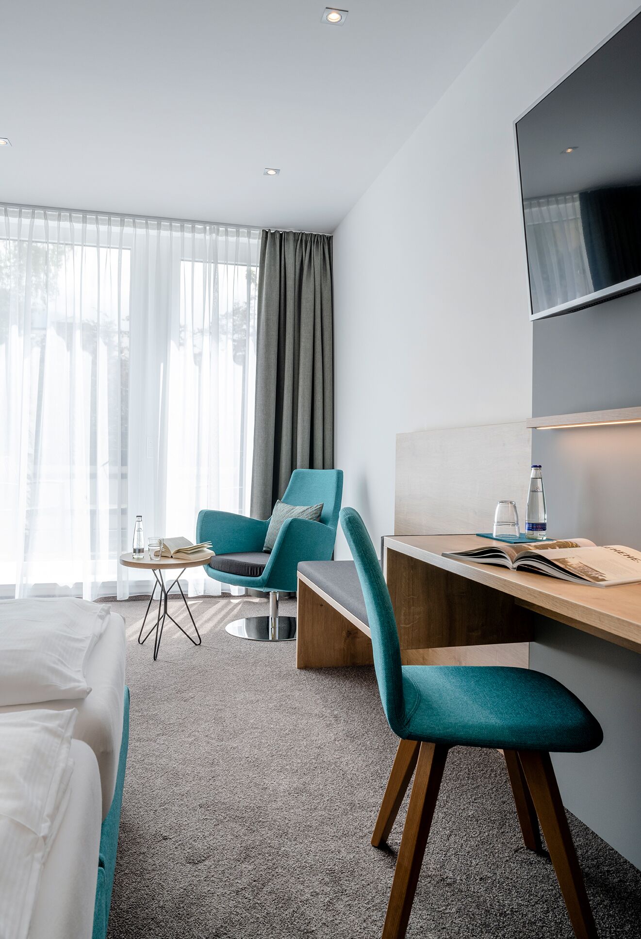 A hotel room with a bed, a desk, two chairs, a TV, and a window with curtains.