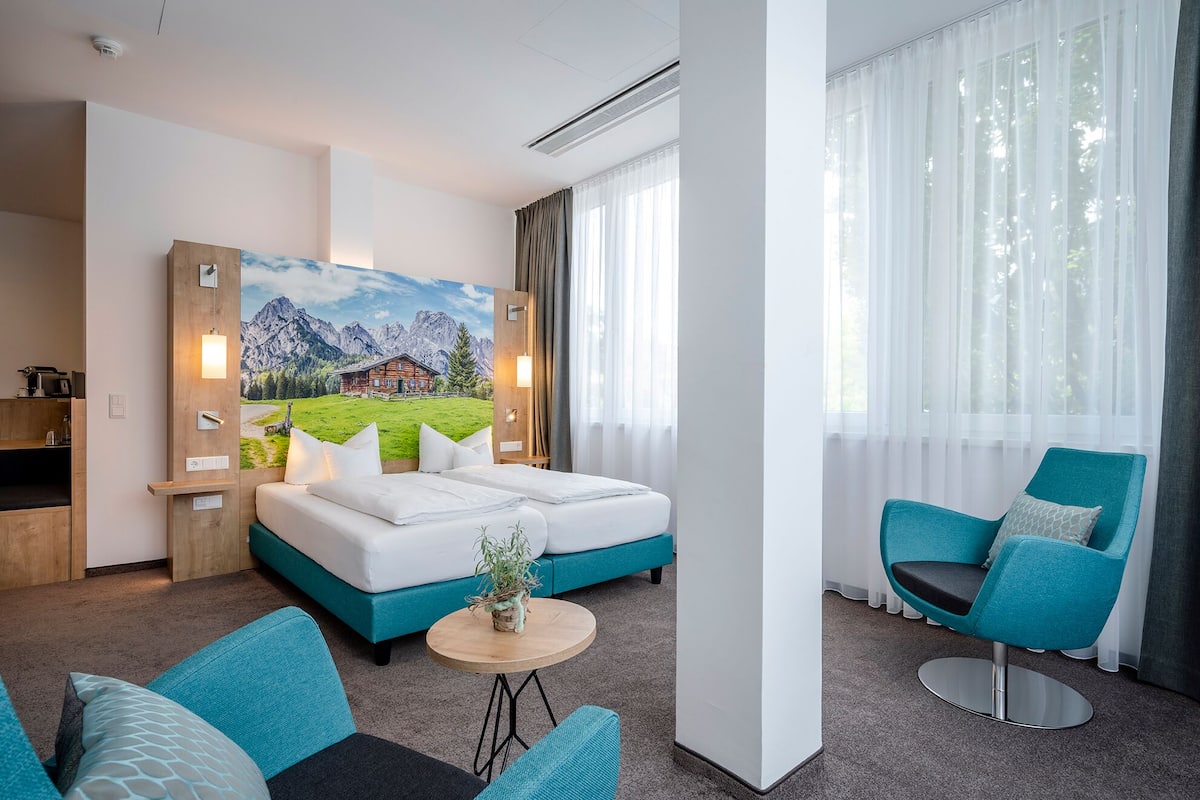 A hotel room with a bed, a desk, a chair, and a painting of a mountain landscape.