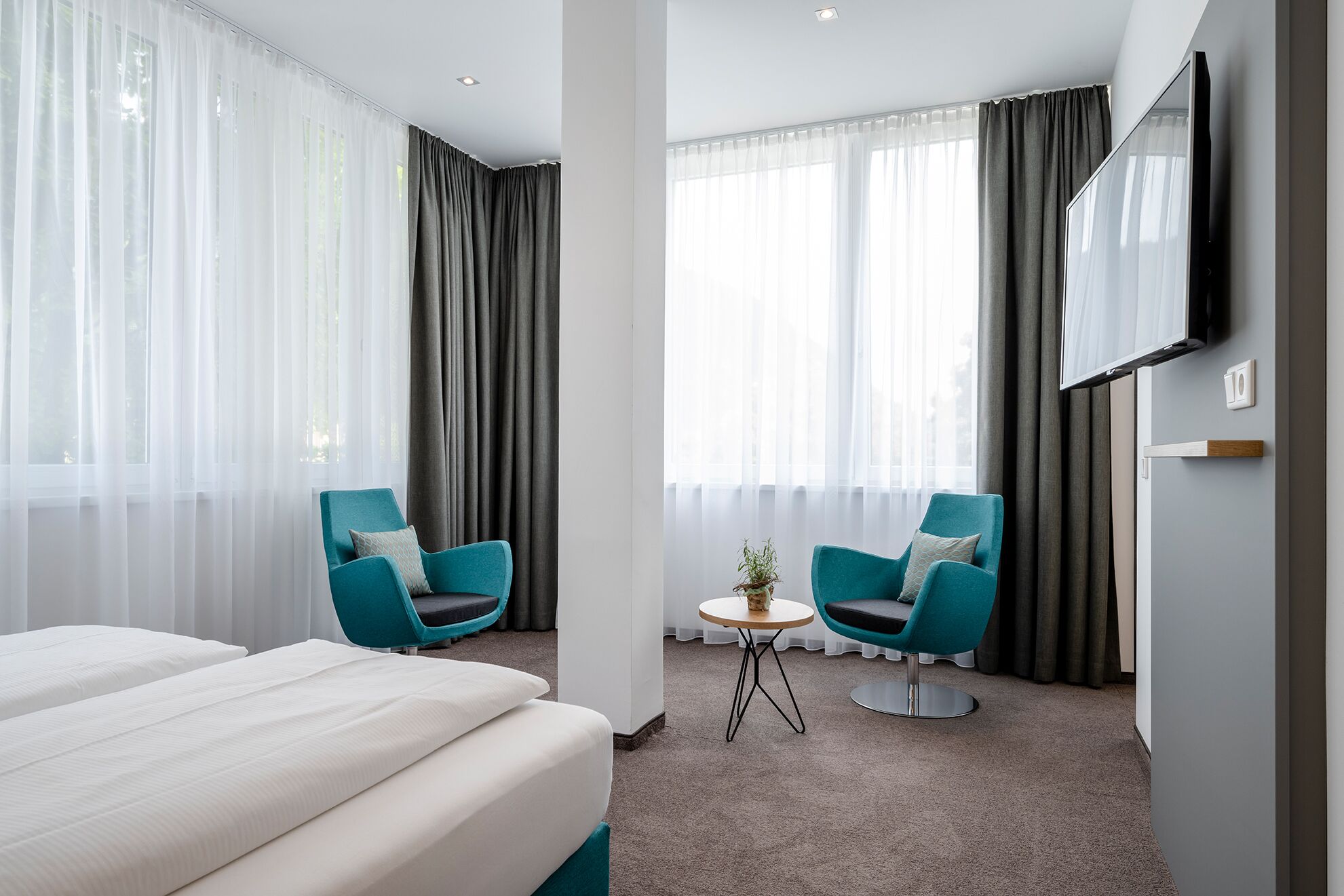Una habitación de hotel con una cama, dos sillones verde mar, una mesita redonda con una planta y un televisor en la pared.